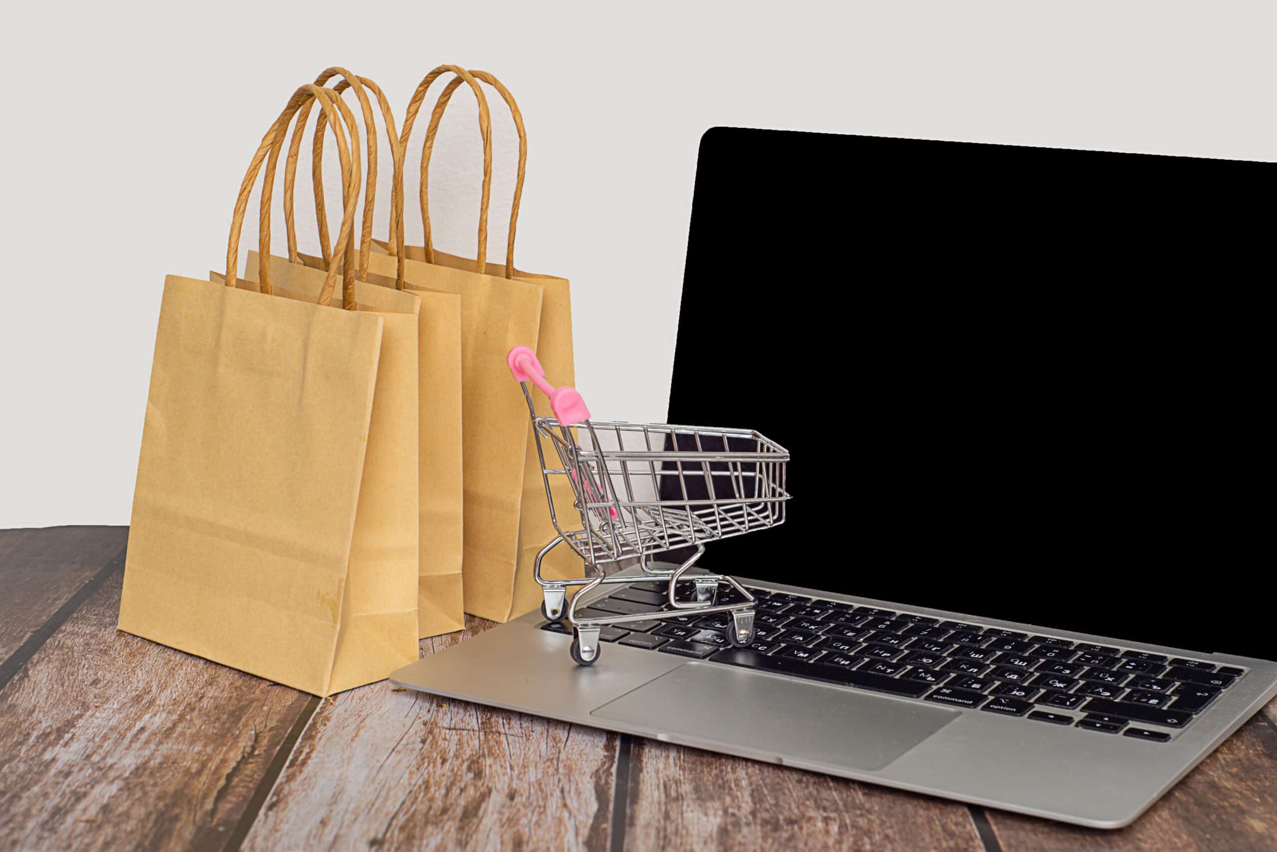 Einkaufswagen auf einem Laptop mit Papiertüten im Hintergrund.